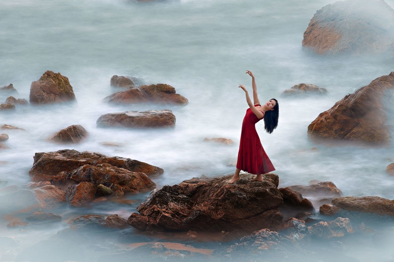 water, rock, rock - object, one person, clothing, sea, solid, standing, young adult, fashion, full length, nature, dress, adult, beauty in nature, motion, women, land, human arm, outdoors, arms raised, flowing, hairstyle