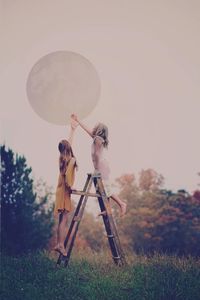 Full length of girls flying large bubble while standing on ladder against clear sky at park