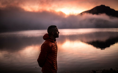 Side view of lonely man in outerwear looking away thoughtfully while relaxing near calm lake water reflecting mountains and cloudy sky at sunset time