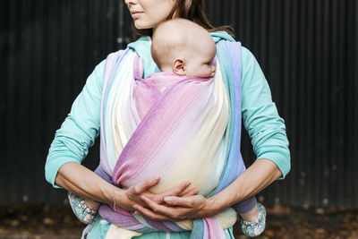 Midsection mother carrying baby wrapped in blanket standing against metal wall