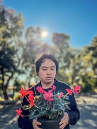 Portrait of young asian man holding red flowering cyclamen potted plant against trees and blue sky