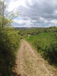 Dirt road passing through field against cloudy sky