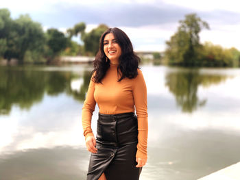 Portrait of smiling young woman standing by lake against sky