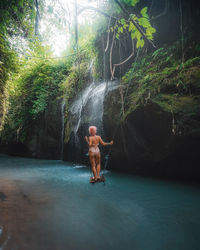Full length rear view of sensual woman standing on rope swing against waterfall in forest