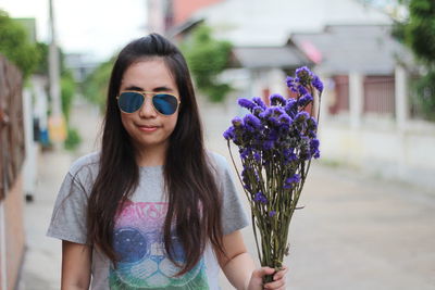 Portrait of woman wearing sunglasses while holding purple flowers on footpath