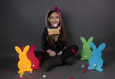 Portrait of smiling girl with eater bunny and pom pom in basket against gray background