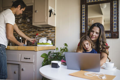 Mother and daughter using laptop while father working in kitchen