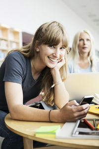 Portrait of smiling young woman holding mobile phone at desk in university library