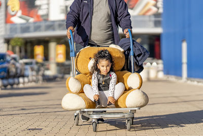 Cute girl sitting with teddy bear in luggage cart on footpath