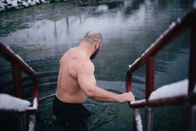 Rear view of shirtless man moving in lake during snowfall in winter