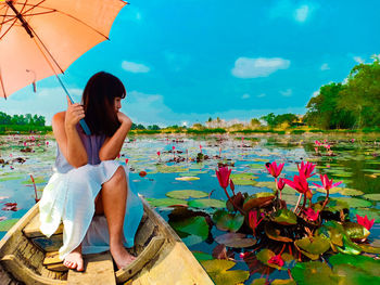 Young woman with umbrella sitting in boat on lake against sky