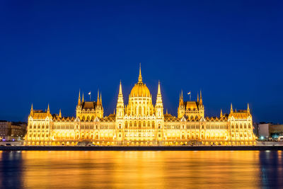 Illuminated hungarian parliament building by danube river against blue sky