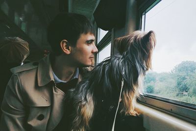 Young man with hairy dog looking through window in train