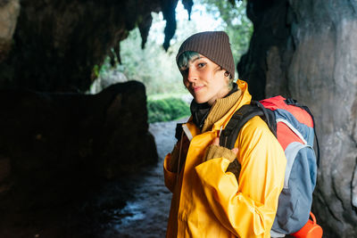 Positive female traveler with backpack and in outerwear standing in rocky arched passage near cobijero beach and looking at camera while enjoying autumn vacation in asturias