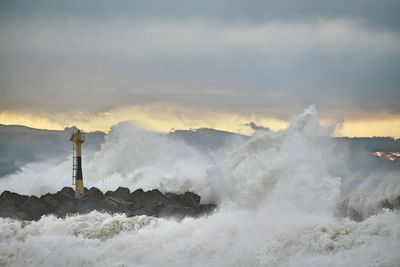 Waves splashing by lighthouse against cloudy sky during sunset