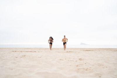 Ground level of diverse sportswoman and sportsman running on sandy shore during fitness training in summer