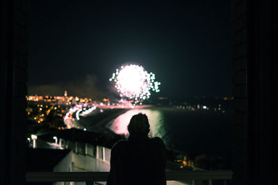 Rear view of silhouette woman looking at firework and illuminated cityscape against clear sky