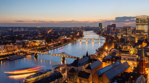 Aerial view of illuminated buildings by river against sky during sunset