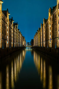 View of illuminated buildings by river against clear blue sky