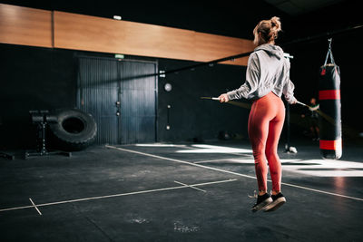 Back view of strong unrecognizable female fighter in sportswear exercising with skipping rope while working out in modern boxing club