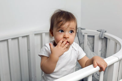 One year old child in white clothes standing in a white round bed in his nursery