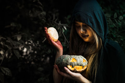 Woman in witch costume holding illuminated jack o lantern by plants during night