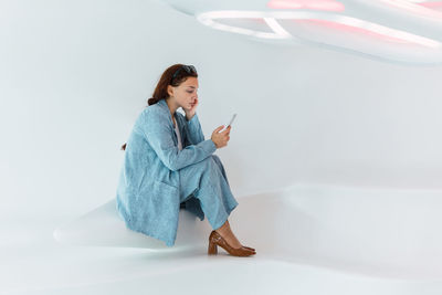 Side view of full body of content female in elegant wear scrolling social media on cellphone while sitting in light modern room with white wall