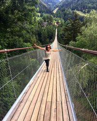 Woman with arms outstretched walking on footbridge against trees