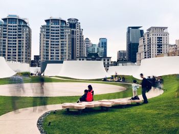 People sitting in park by buildings against sky in city