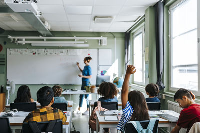 Rear view of girl with hand raised sitting in classroom while female teacher teaching at school