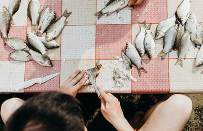 A young man cleans fish from scales while sitting at a table.