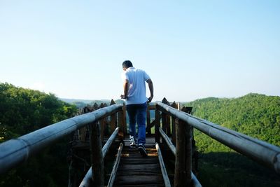 Rear view of man walking on footbridge against clear sky