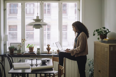 Side view of female construction engineer using laptop while standing with one leg on chair near table at home