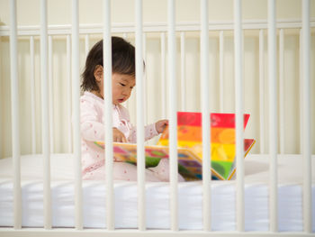 Baby girl reading book while sitting in crib at home
