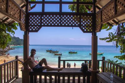 Side view of shirtless young man sitting on bench against sea at tourist resort