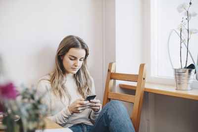 Young woman using mobile phone sitting on chair by window at dorm