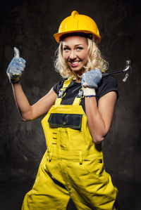 Portrait of smiling female worker showing thumbs up while standing against wall