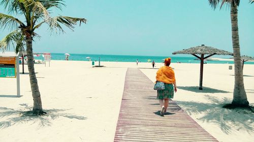 Full length rear view of woman walking on boardwalk at beach against clear blue sky