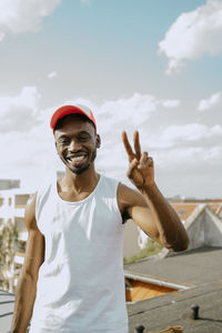 Portrait of young man wearing cap gesturing peace sign against sky on sunny day