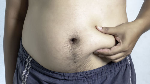 Midsection of shirtless man holding belly while standing against white background