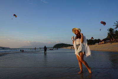 Happy woman having drink while standing at beach against sky