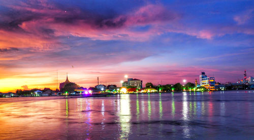 Illuminated buildings by river against sky at sunset