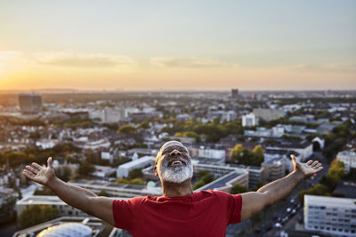 Happy bearded man with arms outstretched while standing on building terrace in city during sunset