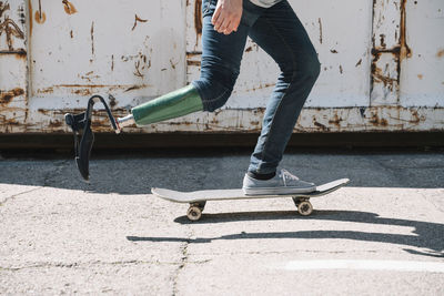 Side view of young guy with leg prosthesis pushing from ground and riding skateboard against metal container on city street