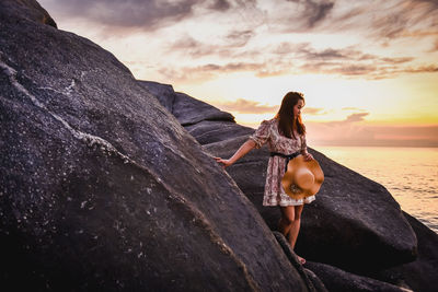 Woman standing on rock by sea against sky during sunset