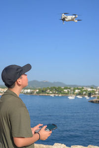 Happy teen boy, flying drone on mediterranean coast against blue sky during sunny day spain,vertical