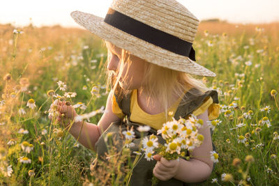 A little blonde girl in a straw hat walks in a field with a bouquet of daisies. 