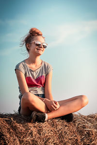 Carefree teenager wearing sunglasses sitting on hay against sky