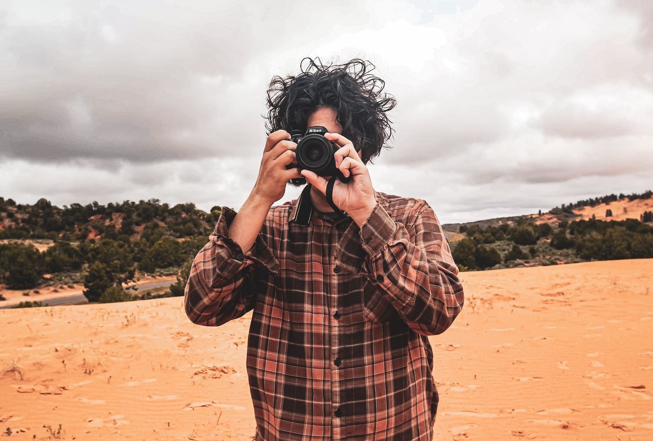 one person, camera - photographic equipment, photography themes, photographing, activity, technology, standing, leisure activity, cloud - sky, real people, sky, occupation, casual clothing, photographer, waist up, men, young adult, holding, nature, outdoors, digital camera, slr camera