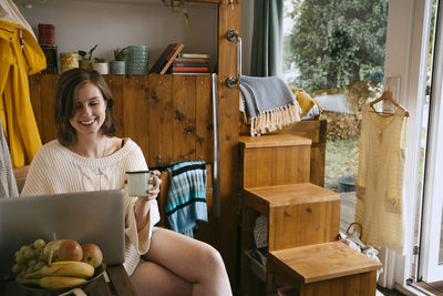 Smiling young woman using laptop holding coffee cup while sitting at home